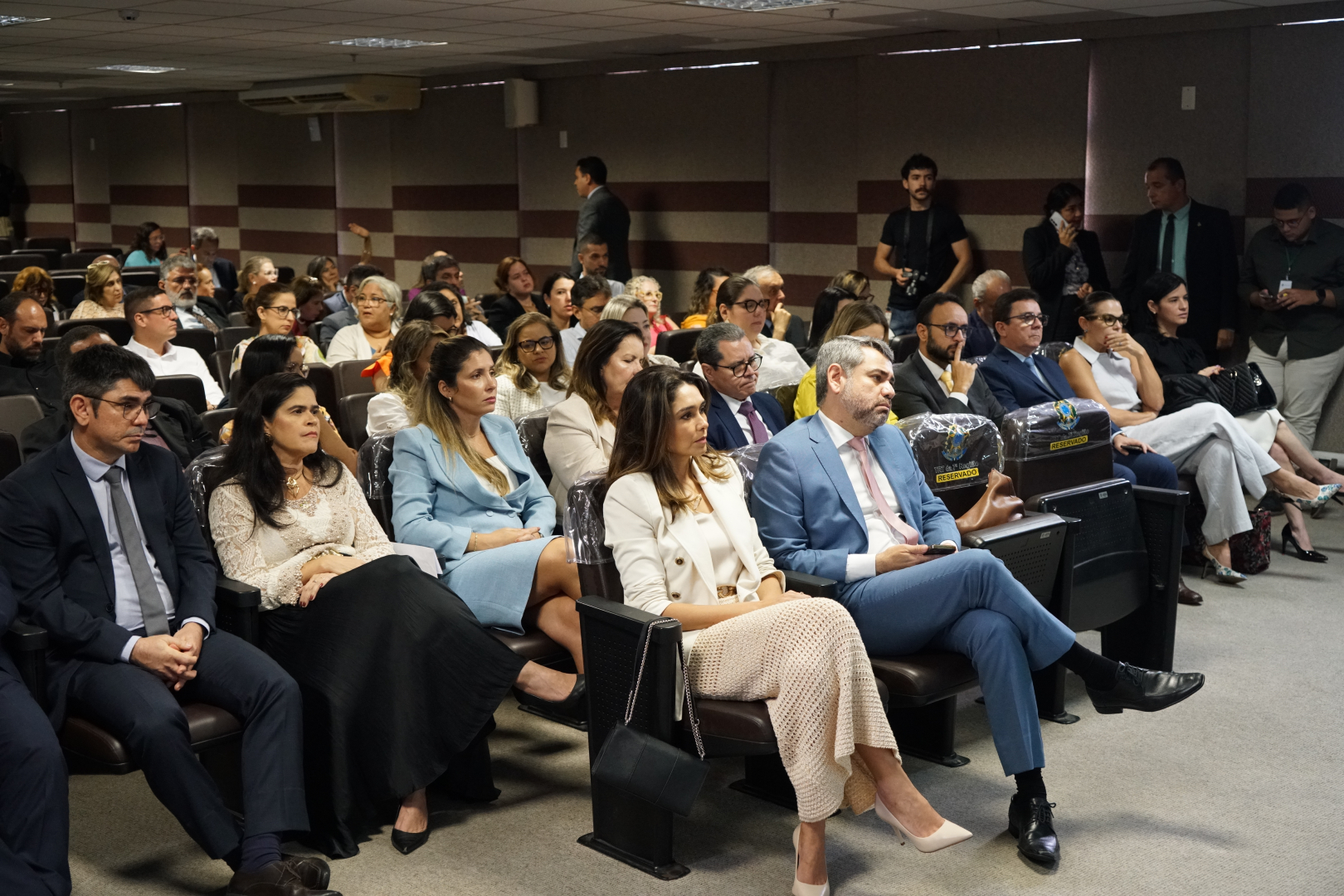 Um público diverso está sentado em cadeiras de um auditório, observando o que acontece à frente. As pessoas, em sua maioria, estão em trajes sociais, algumas atentas e outras conversando. O ambiente tem paredes em tons de marrom e teto com painéis e iluminação embutida.